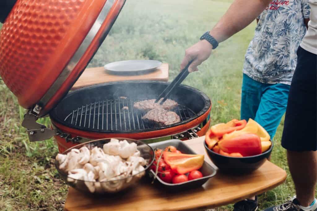 Cooking meat on a kamado grill and a table with sliced mushrooms and vegetables.