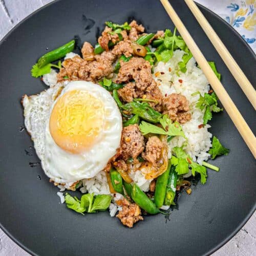 Sizzling Thai Pork & Green Beans topped with egg on a black plate.