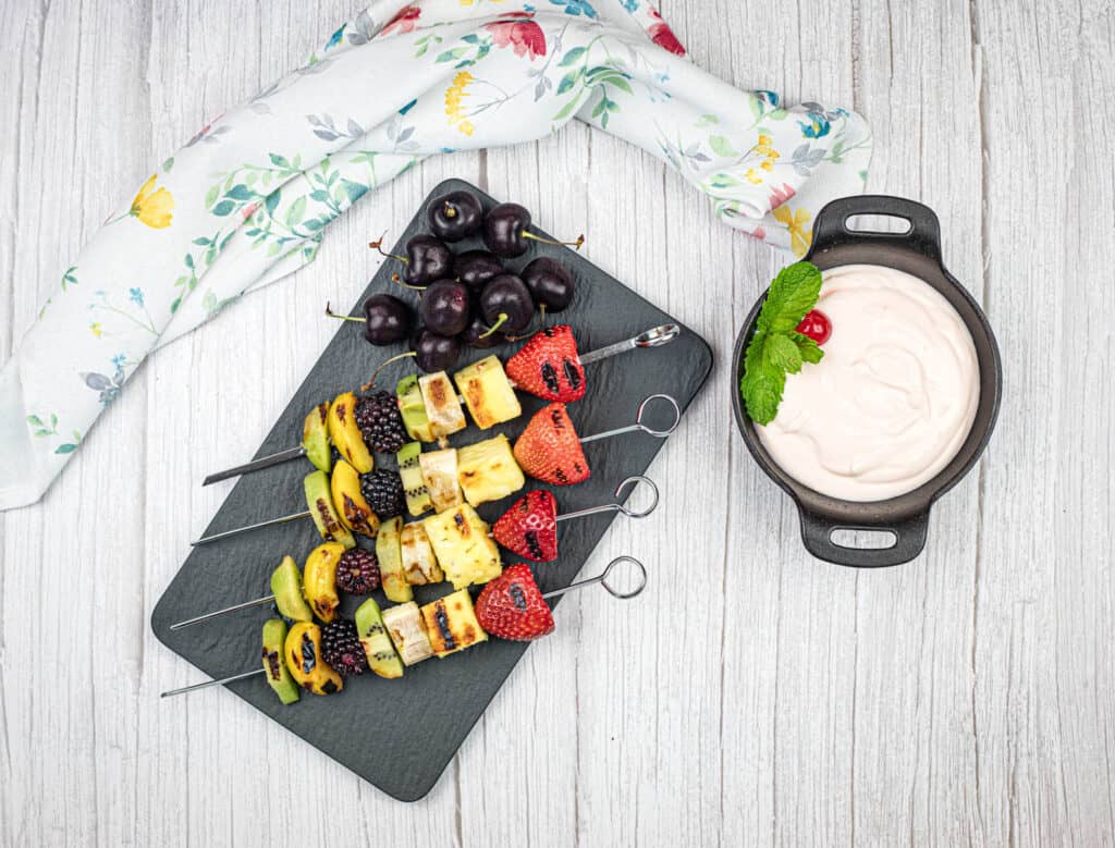 Top-down shot of Four pieces of grilled fruit skewers on a slate plate with a bowl of cherry dip.