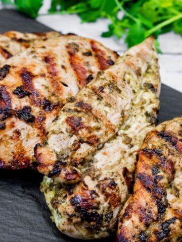 A close-up grilled chicken on a black stone-like board with cilantro leaves in the background.