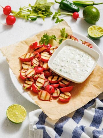 Plate of grilled radishes with charred grill marks served on parchment paper, accompanied by a creamy jalapeño dip garnished with cilantro, surrounded by fresh ingredients including lime, radishes, and jalapeños.