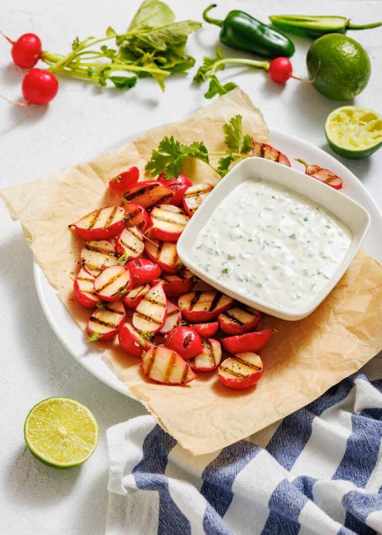 Plate of grilled radishes with charred grill marks served on parchment paper, accompanied by a creamy jalapeño dip garnished with cilantro, surrounded by fresh ingredients including lime, radishes, and jalapeños.