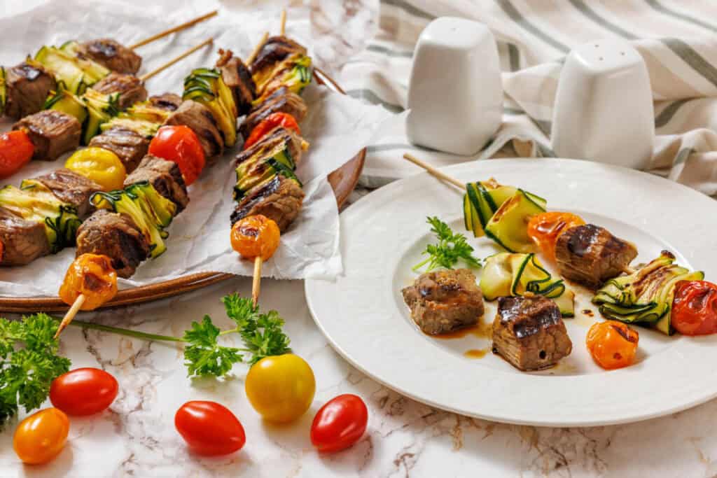 A white marble surface is covered with grilled steak and veggie skewers. On the left, a brown platter holds several skewers, some arranged neatly and some slightly askew, all featuring browned steak, grilled zucchini ribbons, and blistered red and yellow cherry tomatoes.