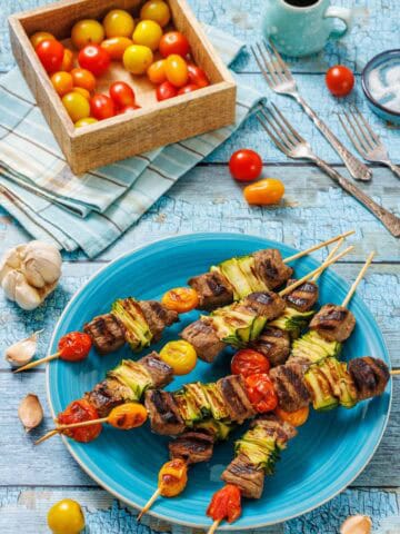 A vibrant blue textured surface serves as the backdrop for a close-up, overhead shot of grilled steak and veggie skewers. A large blue plate in the foreground holds five skewers, each featuring cubes of browned steak, bright green grilled zucchini ribbons, and blistered red and yellow cherry tomatoes.