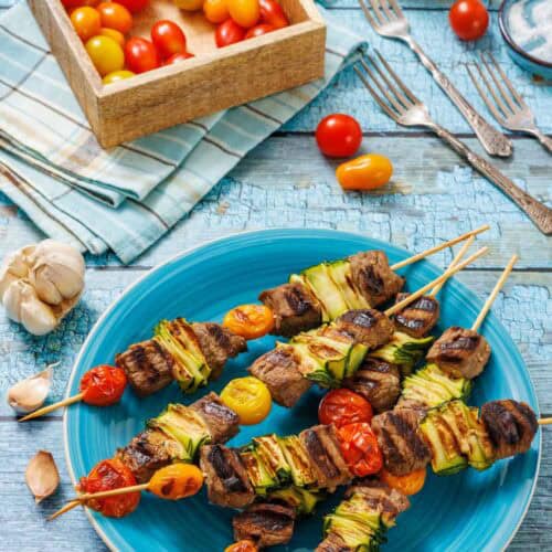 A vibrant blue textured surface serves as the backdrop for a close-up, overhead shot of grilled steak and veggie skewers. A large blue plate in the foreground holds five skewers, each featuring cubes of browned steak, bright green grilled zucchini ribbons, and blistered red and yellow cherry tomatoes.