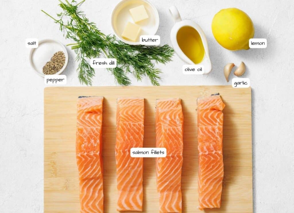 An overhead view of a white countertop displaying labeled ingredients for Lemon Dill Salmon Foil Packets, including four salmon fillets, salt, pepper, fresh dill, butter, olive oil, a whole lemon, and garlic cloves.