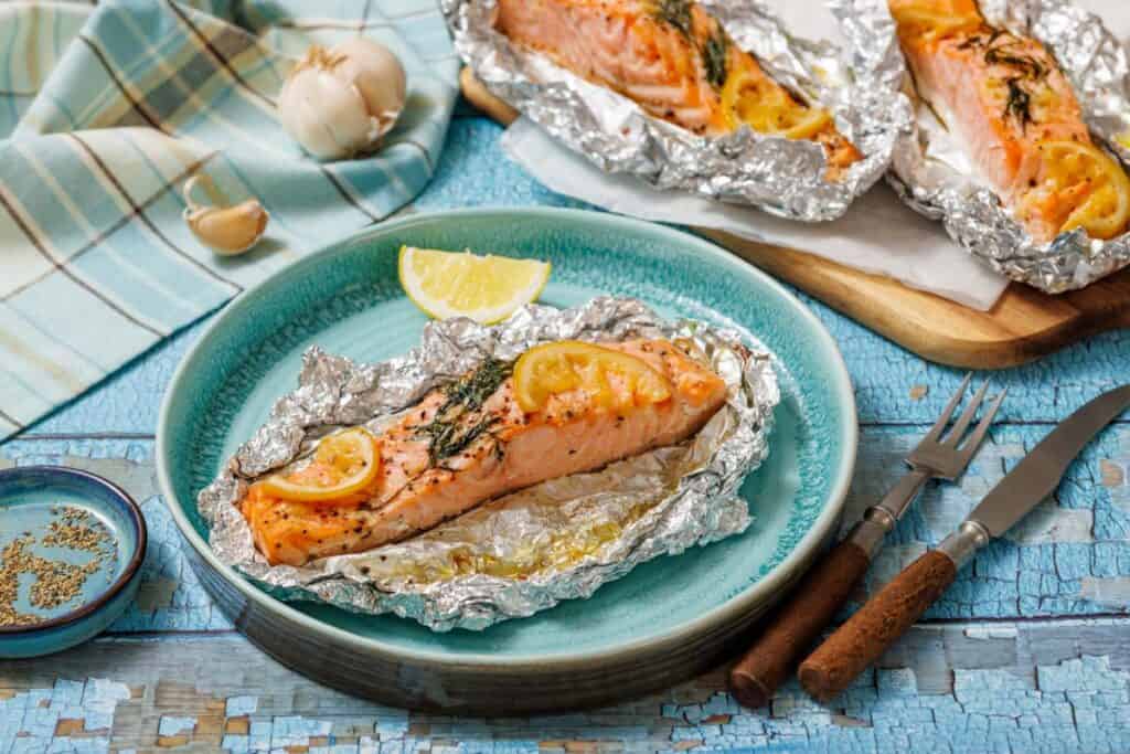 A cooked salmon fillet seasoned with lemon slices and dill, still in its foil packet, rests on a teal plate with a lemon wedge, fork, knife, and a small bowl of spices on a blue wooden surface.