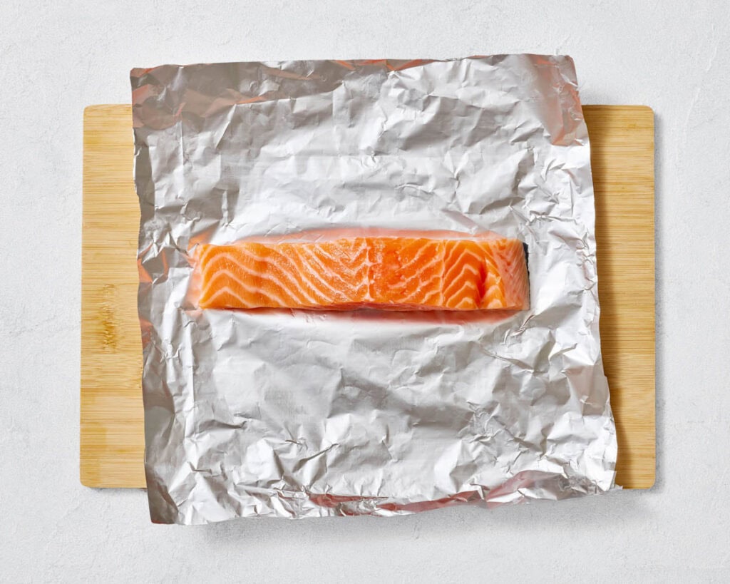 An overhead shot of a single, large, orange-pink raw salmon fillet with distinct white fat lines, placed on a square sheet of crinkled aluminum foil, which rests on a light wooden cutting board.