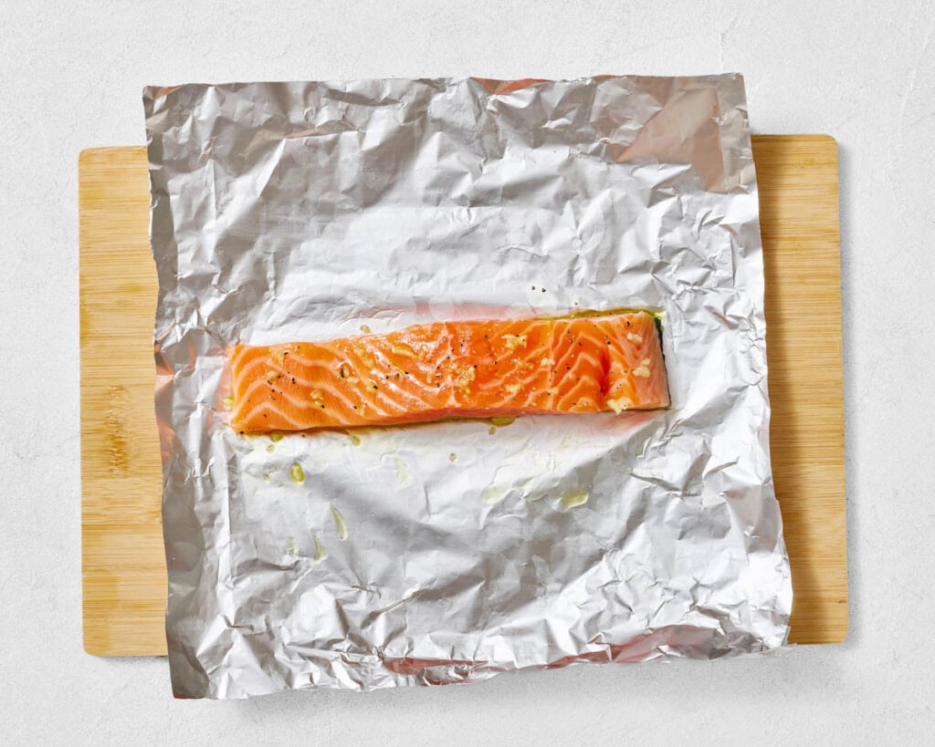 An overhead shot of a single, large, orange-pink raw salmon fillet, sprinkled with minced garlic and black pepper, resting on a sheet of crinkled aluminum foil on a wooden cutting board.