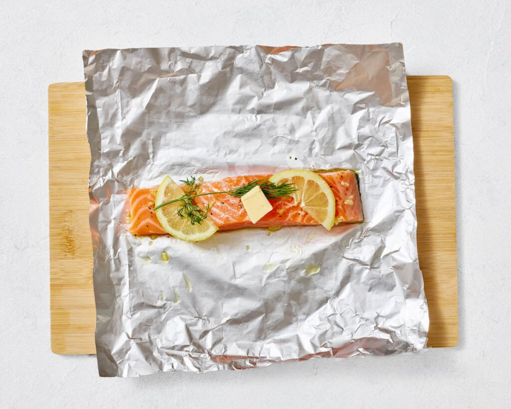 An overhead shot of a raw salmon fillet on crinkled aluminum foil, topped with two lemon slices, a sprig of fresh dill, and a pat of butter, all on a wooden cutting board.