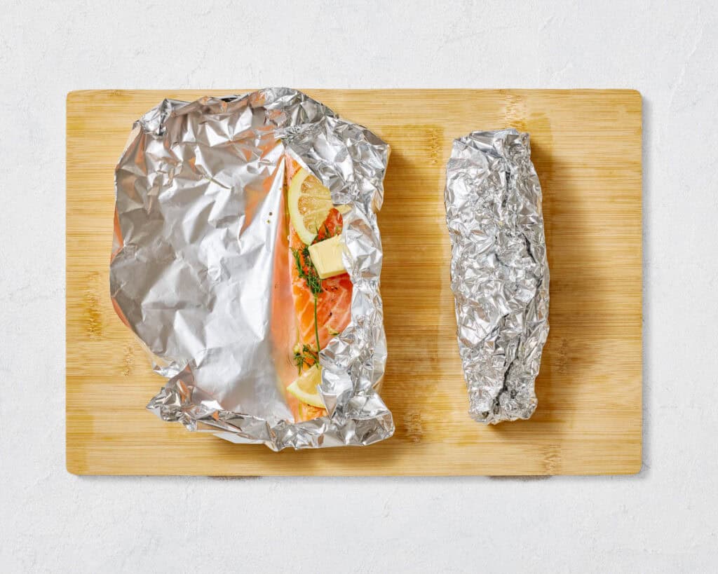 An overhead shot of a raw salmon fillet in an open, crinkled aluminum foil packet topped with lemon, dill, and butter, next to a fully sealed foil packet containing another salmon fillet, all on a wooden cutting board.