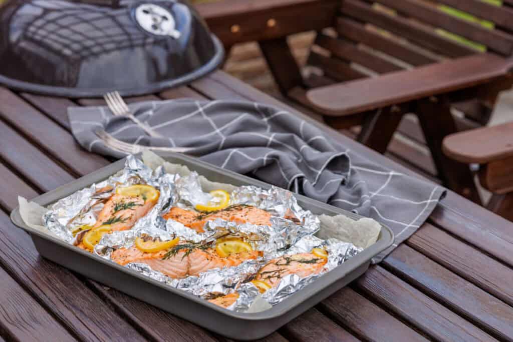 A dark gray baking pan filled with several cooked Lemon-Dill Salmon fillets, still in their open aluminum foil packets with visible lemon slices and dill, rests on a dark wooden slatted outdoor table, with a blurred grill and a gray plaid napkin in the background.