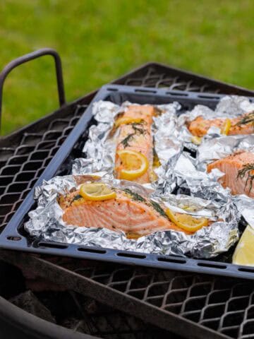 Several large salmon fillets are arranged on individual foil sheets on a black baking tray, seasoned with lemon slices and fresh herbs, and placed on a metal grill grate outdoors.