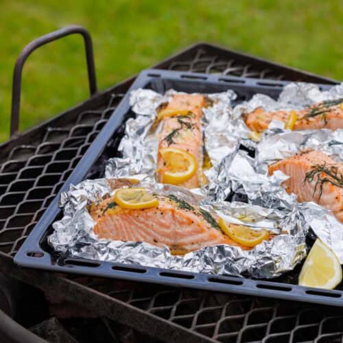 Several large salmon fillets are arranged on individual foil sheets on a black baking tray, seasoned with lemon slices and fresh herbs, and placed on a metal grill grate outdoors.