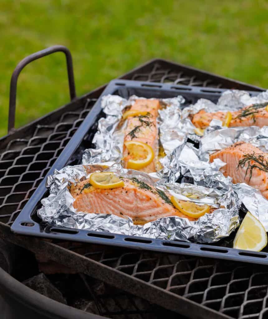 Several large salmon fillets are arranged on individual foil sheets on a black baking tray, seasoned with lemon slices and fresh herbs, and placed on a metal grill grate outdoors.