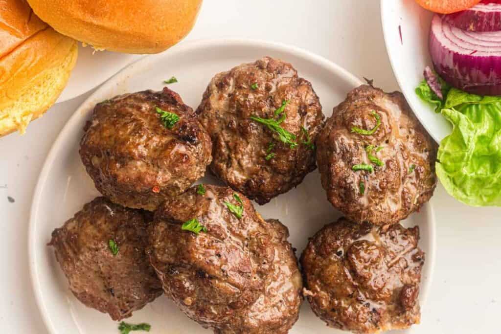 Seven pieces of grilled beef burgers on a round plate beside buns and vegetables.